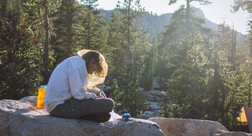 a person wearing a mosquito net sits on a rock while writing in a journal on an outward bound course for bipoc